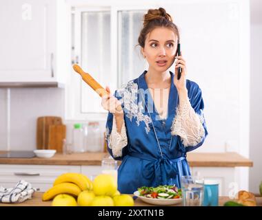 Femme insatisfaite avec la goupille dans les mains parlant au téléphone dans kichen Banque D'Images