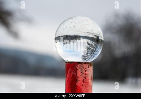 Boule de verre couchée sur un poteau en métal rouge rouillé sur une froide journée d'hiver dans la nature, avec des reflets d'un paysage enneigé dans le verre Banque D'Images