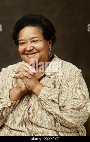 Portrait moyen de femme afro-américaine âgée souriante dans des vêtements beiges se pliant les mains de manière heureuse tout en regardant loin sur fond brun dans le studio Banque D'Images