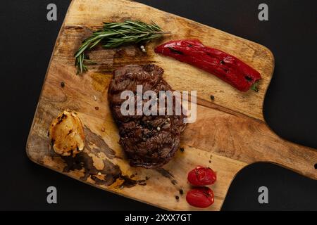 Vue de dessus de Rib Eye Black Angus grillé servi avec du poivre et des herbes sur une planche de bois sur fond sombre. Banque D'Images