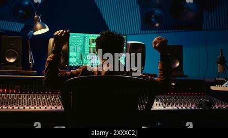 Producteur de musique afro-américain vibing sur son nouveau morceau en studio professionnel, mixant et mastering des airs en salle de contrôle. Technicien appuyant sur les boutons et les curseurs du panneau de la table d'harmonie. Caméra B. Banque D'Images