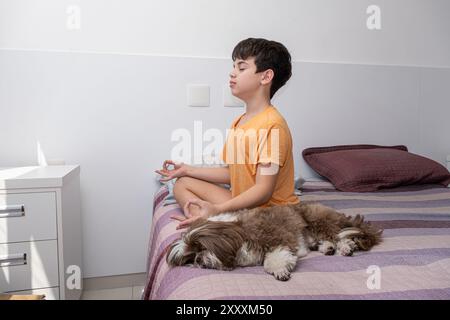 Garçon brésilien de 11 ans assis sur son lit, pratiquant le yoga à côté de son chien 1. Banque D'Images