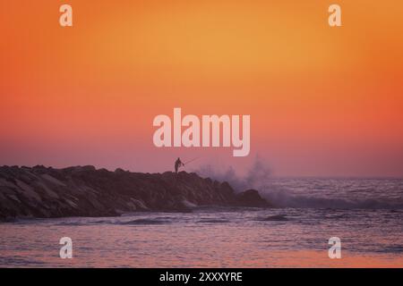 Pêche de silhouette de pêcheur dans l'océan atlantique avec des vagues s'écrasant sur un quai en pierre au coucher du soleil, au Portugal Banque D'Images