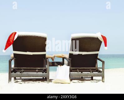 Carte de Noël ou fond, couple dans des chaises longues avec chapeaux de Père Noël debout sur une belle plage tropicale avec sable blanc et eau turquoise. Concept Banque D'Images