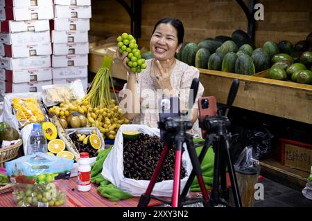 Une joyeuse vendeuse de fruits diffusant en direct ses produits, y compris des cerises, des oranges et des avocats, sur un marché de Phnom Penh, au Cambodge. Banque D'Images