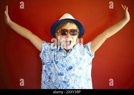 Très emothional enfant porter un chapeau et des lunettes de soleil sur fond rouge. Copie espace, lumière du jour Banque D'Images