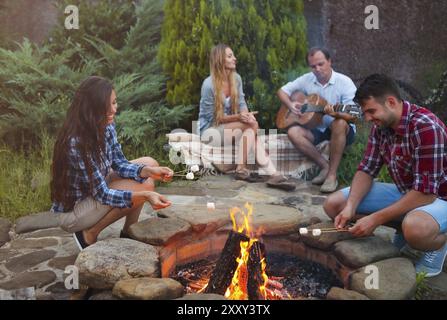 Compagnie des jeunes gens tenant des bâtons avec des guimauves au-dessus du feu de camp en soirée. Maison de vacances et d'amitié concep Banque D'Images