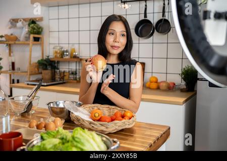 Une blogueuse gastronomique asiatique attrayante ou créatrice de contenu cuisine devant la caméra, parle et montre ses ingrédients pour un menu spécial W. Banque D'Images