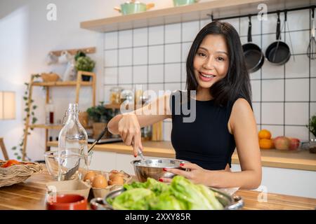 Une femme asiatique magnifique fouette le mélange de crêpes dans un bol à la table de la cuisine tout en préparant le petit déjeuner. petit déjeuner, nourriture saine, recette, lif domestique Banque D'Images