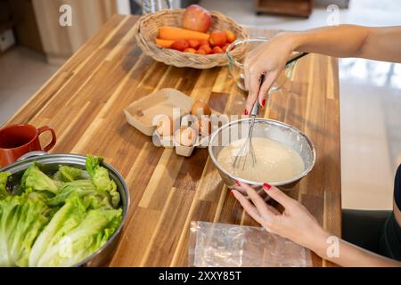 Une image en gros plan d'une femme fouette la pâte à crêpes mélangée dans un bol à la table de la cuisine tout en préparant le petit déjeuner. petit déjeuner, nourriture saine, recette, Banque D'Images