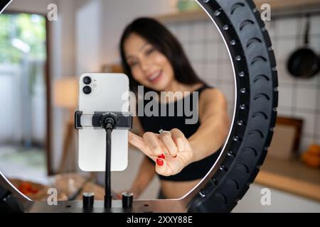 Une image en gros plan d’une influenceuse asiatique ajustant la caméra sur son smartphone, se préparant à aller en direct sur les réseaux sociaux tout en cuisinant dans le kitch Banque D'Images