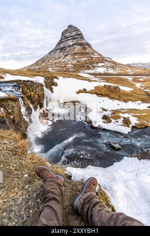 POV d'un homme assis sur la cascade avec de la neige sur la célèbre montagne Kirkjufell en hiver Islande Banque D'Images