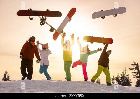 Groupe d'amis heureux snowboarders jettent des snowboards et sautent contre le ciel du coucher du soleil. Concept de station de ski d'hiver Banque D'Images