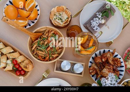 Vue de haut en bas d'une élégante configuration de salle à manger avec des plats méditerranéens comprenant pâtes, poulet, pain assorti, fruits frais et boissons, disposé sur une table en bois avec des fleurs ajoutant du charme Banque D'Images