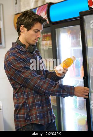 Homme, réfrigérateur et shopping dans la boutique pour jus, soda et décision avec écouteurs, étiquette et lecture. Étudiant, réfrigérateur et choix dans un supermarché ou Banque D'Images