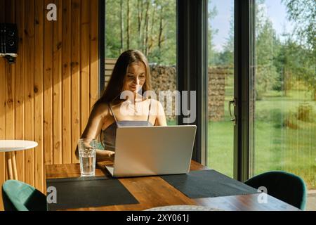 Femme joyeuse assise à une table en bois dans une maison de campagne engrossée dans le travail sur un ordinateur portable Banque D'Images