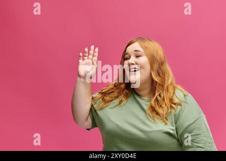 Une femme aux longs cheveux rouges sourit et agite bonjour sur un fond rose. Banque D'Images