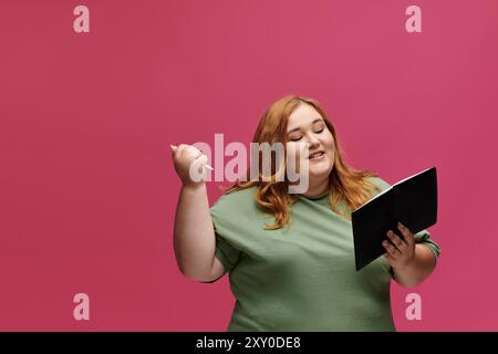 Une femme en chemise verte lit un livre, tenant un stylo dans son autre main, souriant comme elle le fait. Banque D'Images