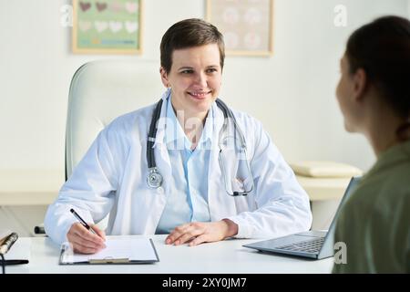 Portrait d'une femme médecin senior portant un manteau blanc et un stéthoscope, s'engageant dans une conversation amicale avec le patient tout en prenant des notes dans un cadre de bureau avec des tableaux médicaux en arrière-plan Banque D'Images