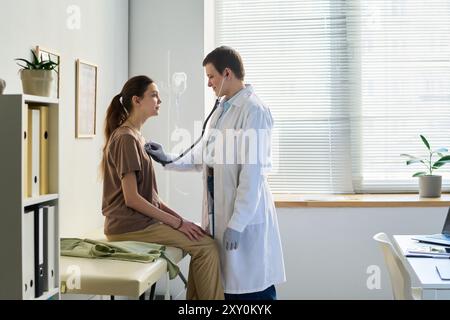 Médecin effectuant un examen physique à l'aide d'un stéthoscope sur le patient assis dans le cabinet médical avec filtrage de la lumière naturelle à travers la fenêtre et divers outils médicaux visibles Banque D'Images