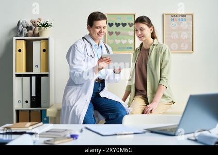 Médecin consultant le patient dans le cabinet médical lumineux, discutant des résultats affichés sur tablette tout en souriant. Étagères avec classeurs et affiches santé visibles en arrière-plan Banque D'Images