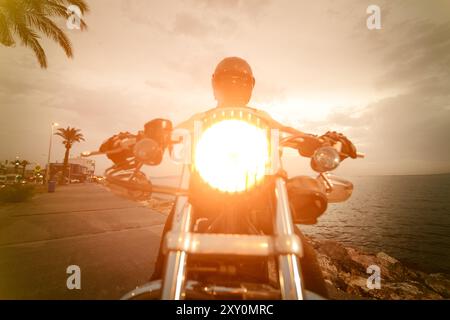 Un motard, entièrement équipé avec casque, lunettes et couvre-visage, est assis sur une moto chopper avec le phare allumé, prêt à rouler dans le coucher du soleil par th Banque D'Images