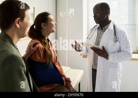 Couple souriant parlant au médecin afro-américain tenant la tablette dans le cabinet médical avec le stand IV en arrière-plan. Médecin donnant des conseils et expliquant les détails Banque D'Images