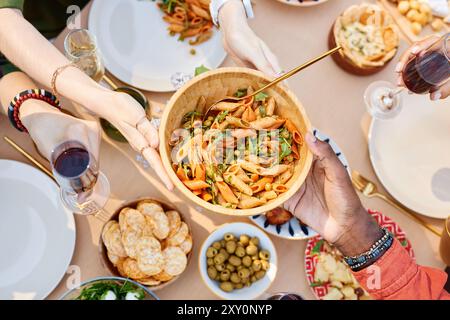 Mains passant un bol de pâtes à une table avec divers plats, vu d'en haut. Groupe diversifié appréciant un repas de cuisine mixte, avec des pâtes, des olives et des craquelins Banque D'Images