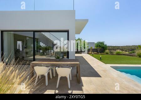 Maison contemporaine affichant un design élégant et moderne avec de grandes fenêtres L'espace extérieur dispose d'une table à manger en bois, de chaises élégantes et d'un pris Banque D'Images