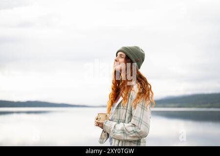 Une jeune femme aux cheveux roux dans un bonnet vert confortable et veste à carreaux appréciant paisiblement une boisson chaude au bord d'un lac tranquille, incarnant l'essence de Banque D'Images