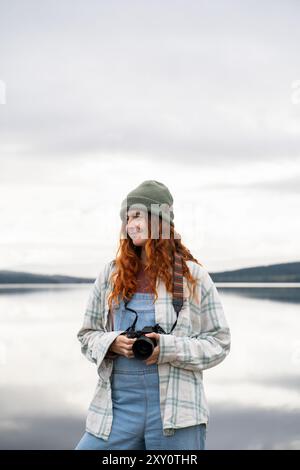 Une jeune femme campeuse avec les cheveux roux bouclés se tient au bord d'un lac tranquille, tenant un appareil photo, vêtue d'une combinaison décontractée en denim, d'une chemise rayée et d'un comfor Banque D'Images