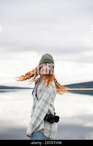 Une jeune femme souriante aux cheveux roux bouclés profite d'un voyage de camping au bord d'un lac serein portant un bonnet vert confortable et une chemise à carreaux, elle tient un appareil photo, Re Banque D'Images