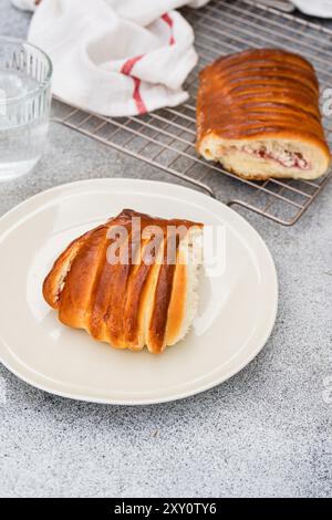 Brioche maison fourrée de cerise et de fromage cottage, servie sur une assiette blanche à côté d'une grille de refroidissement avec un pain entier. Banque D'Images