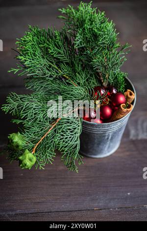Un seau en métal rustique rempli de boules de Noël rouges éclatantes et de bâtons de cannelle, niché au milieu de branches de sapin vert luxuriant, sur un bois sombre Banque D'Images