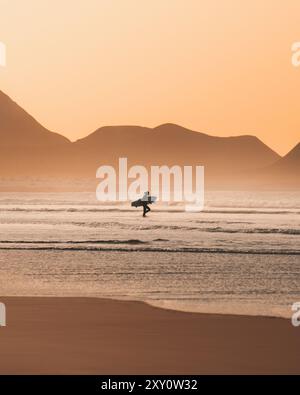 Vue latérale d'un surfeur méconnaissable tenant une planche de surf, marchant vers les vagues de la mer au coucher du soleil, avec des silhouettes de montagne en arrière-plan. Banque D'Images