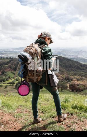 Vue arrière d'une randonneuse méconnaissable admirant de vastes collines verdoyantes tout en utilisant un smartphone. Elle est équipée d'un sac à dos lourdement chargé, w Banque D'Images