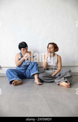 Deux sœurs adultes s'engagent dans un moment décontracté tout en étant assis sur un sol gris contre un mur blanc fumant une cigarette, toutes deux sont habillées en denim décontracté Banque D'Images