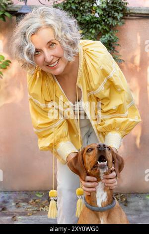 Une femme mature joyeuse aux cheveux gris bouclés sourit et interagit de manière ludique avec son chien brun de taille moyenne dans un jardin ensoleillé. Banque D'Images