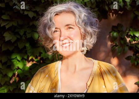 Une femme mature aux cheveux argentés, vêtue d'une robe d'été jaune, sourit chaleureusement tout en se tenant dehors dans un jardin ensoleillé, laisse dans le fond, Banque D'Images