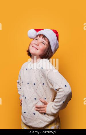 Une jeune fille de preteen joyeuse portant un chapeau de Père Noël regarde vers le haut avec un large sourire, placé sur un fond jaune Uni. Banque D'Images