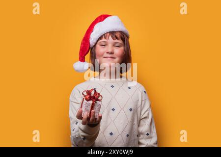Une jeune fille de preteen joyeuse portant un chapeau de Père Noël et un pull confortable sourit doucement tout en tenant un petit cadeau de Noël enveloppé vers la caméra sur un brick Banque D'Images