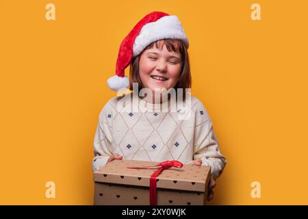 Une jeune fille preteen joyeuse portant un chapeau de Père Noël ferme ses yeux dans le bonheur tout en tenant une boîte cadeau de Noël festive sur un fond jaune vif. Banque D'Images