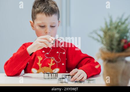 Un jeune garçon portant un pull rouge sur le thème des fêtes façonne méticuleusement les biscuits de Noël à l'aide de couteaux en métal, symbolisant la préparation festive traditionnelle Banque D'Images