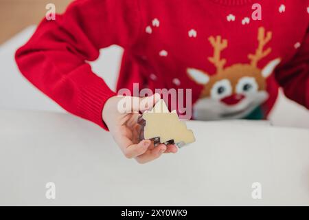 Un enfant dans un pull de Noël rouge tient un emporte-pièce en forme de sapin, soulignant la joie de la cuisson des fêtes Banque D'Images