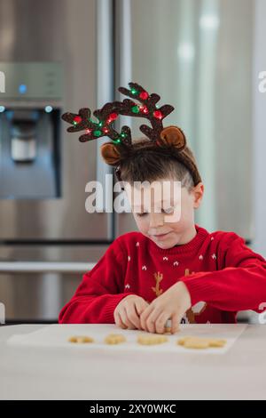 Un jeune garçon avec un bandeau en bois de renne se concentre sur la décoration des biscuits, vêtu d'un pull de Noël rouge vif, incarnant l'esprit festif Banque D'Images