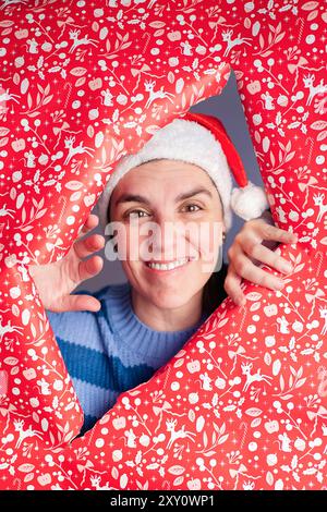 Une femme joyeuse portant un chapeau de Père Noël pairs à travers un papier cadeau rouge de fête déchiré décoré de motifs de Noël blancs, émanant de joyeuses vacances s. Banque D'Images