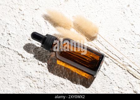 Un flacon compte-gouttes en verre brun repose sur une surface en pierre blanche texturée, accompagné d'une délicate herbe de pampa sèche sous la lumière naturelle du soleil, soulignant l'éco Banque D'Images