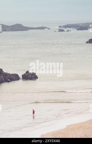 Vue arrière d'une personne méconnaissable debout sur une plage tranquille, face à la mer avec des falaises et des formations rocheuses en arrière-plan. La plage est douce Banque D'Images