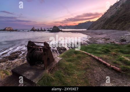 Un lever de soleil tranquille illumine la plage de Gueirua dans les Asturies, avec un treuil rouillé au premier plan et des formations rocheuses saisissantes le long du rivage. Banque D'Images