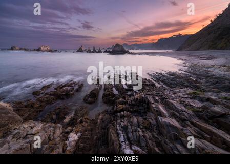 Un lever de soleil serein illumine la plage de Gueirua dans les Asturies, projetant une douce lueur sur les formations rocheuses escarpées et les eaux tranquilles, créant un pittoresque Banque D'Images